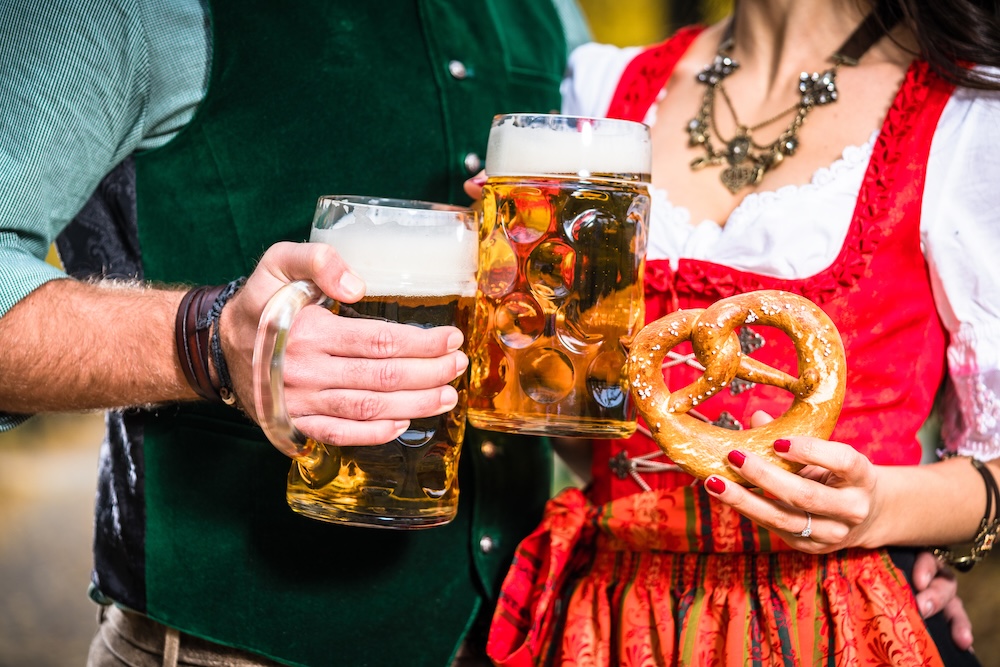A man and woman in traditional Bavarian attire holding glasses of beer and a pretzel